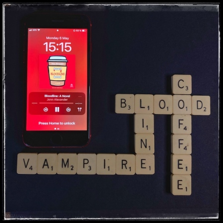 A red iPhone with the cover of Bloodline by Jenn Alexander, narrated by Quinn Riley, on a black background. Next to the phone, Scrabble letters form the words Blood, Line, Vampire, Coffee.
