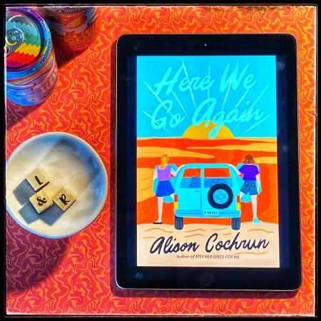 An iPad with the cover of Here We Go Again by Alison Cochrun against a swirly orange backdrop, with a couple of colourful soda cans and the letters L&R in a small bowl.